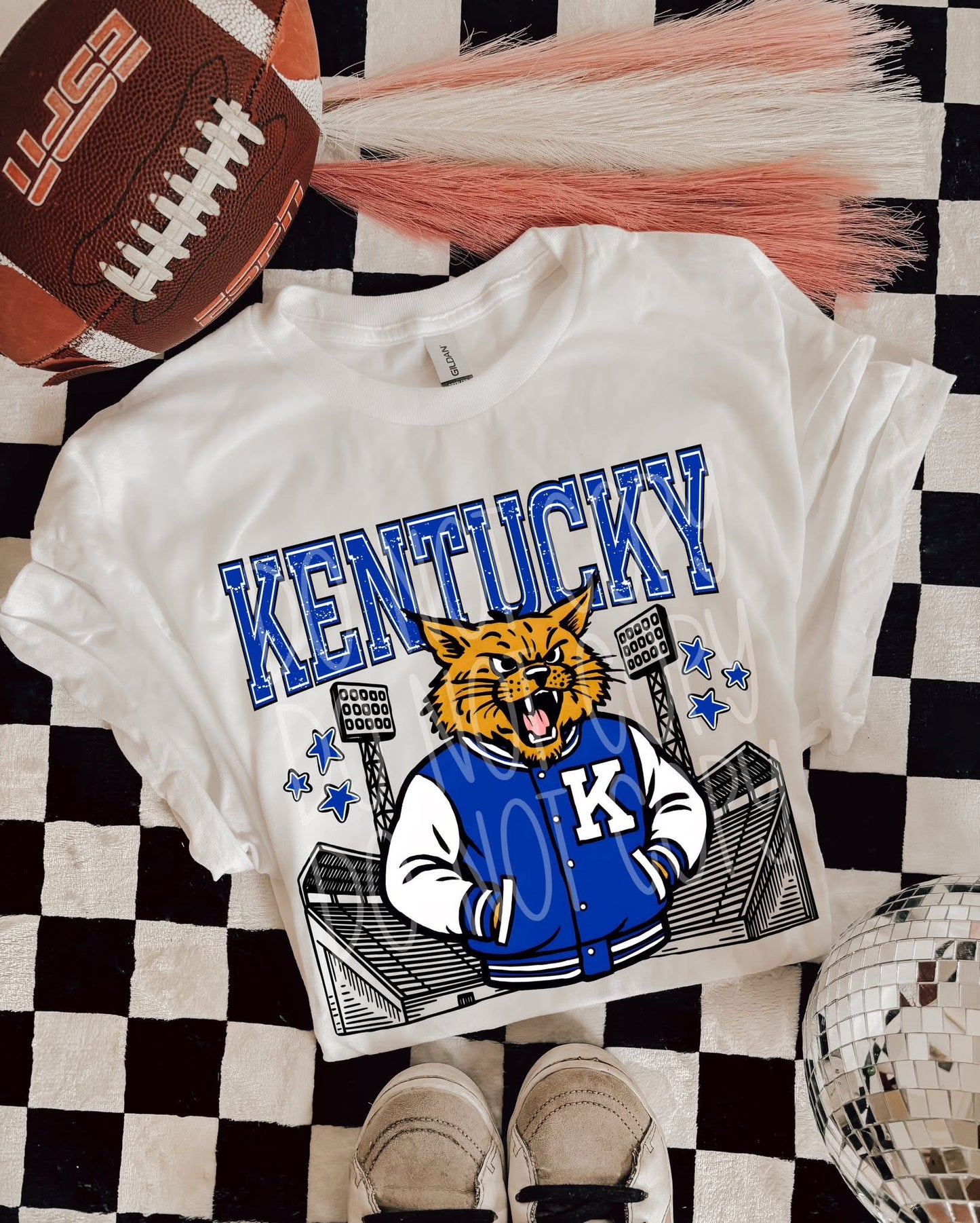 White t-shirt with 'Kentucky' and a mascot design on a checkered surface with a football and pom-poms.